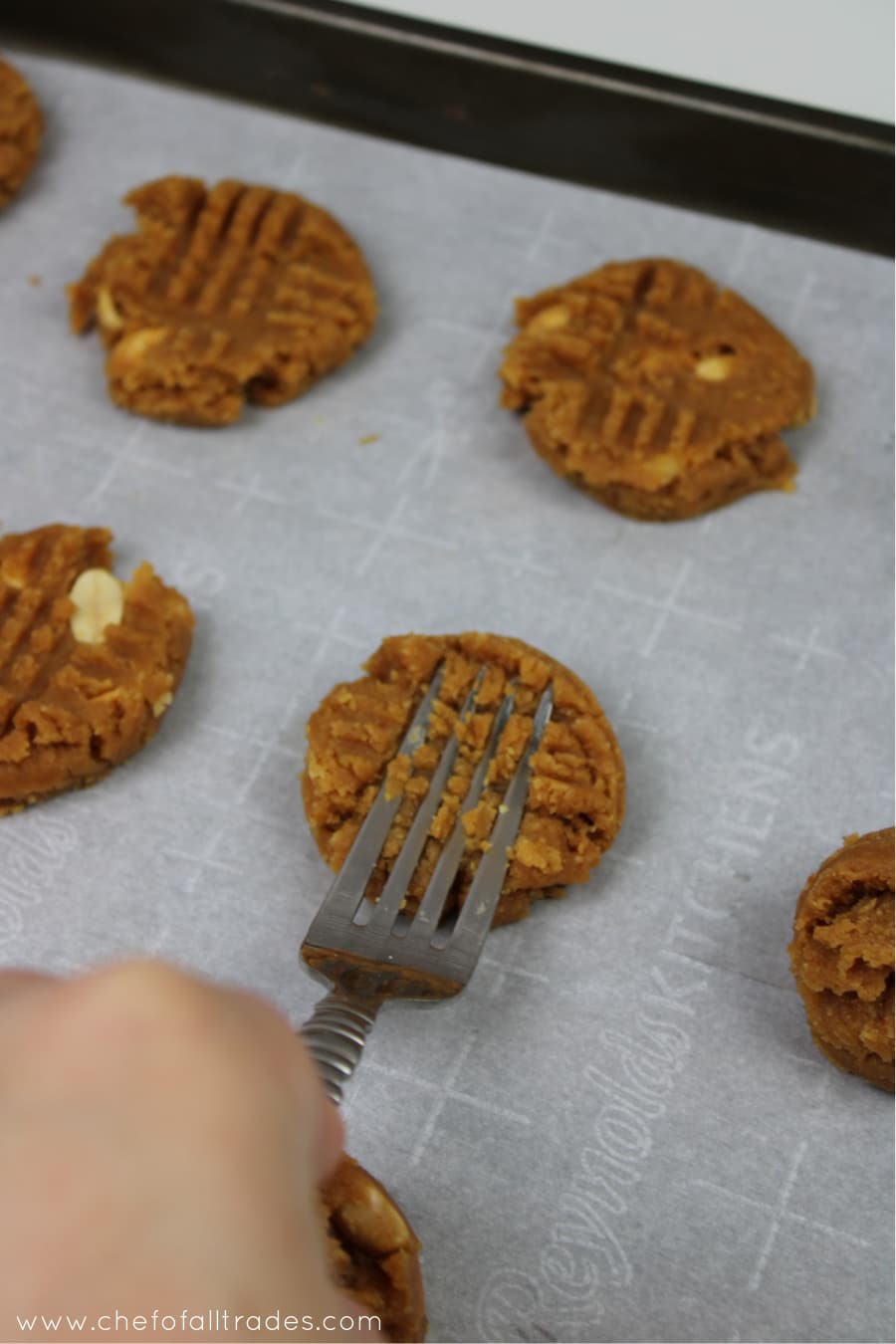 cookie dough scooped onto a parchment lined cookie sheet and being pressed with a fork into cookie shape