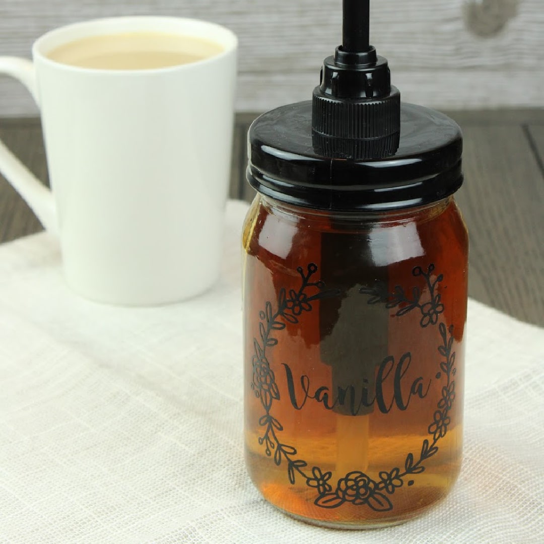 vanilla coffee syrup in a glass jar with a pump attached. "vanilla" label is on the jar with a white cup of coffee in the background.